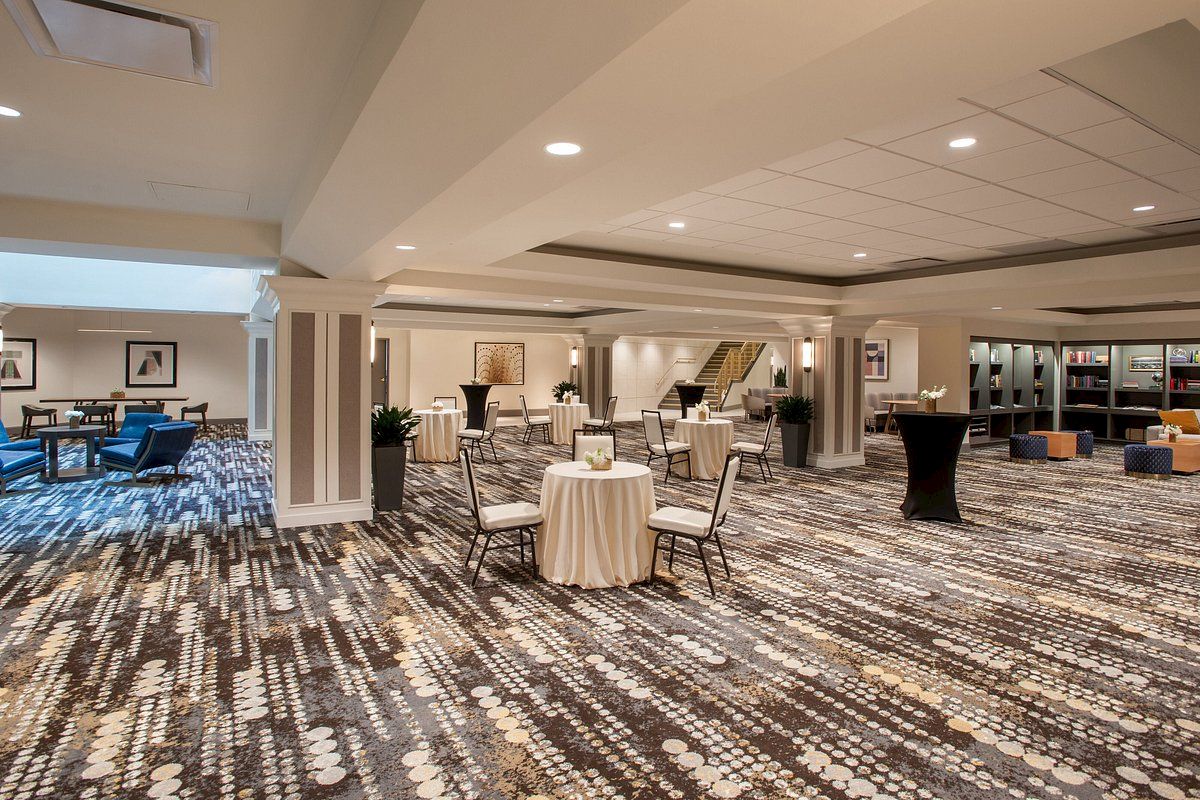 The image shows a modern hotel lobby with patterned carpet, tables with flowers, chairs, and a seating area in the background.