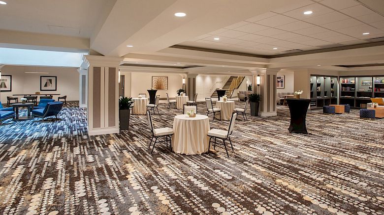 The image shows a modern hotel lobby with patterned carpet, tables with flowers, chairs, and a seating area in the background.