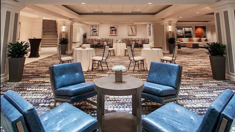 The image shows a modern hotel lobby with a glass ceiling, blue chairs around tables, and a bright, open space with plants.