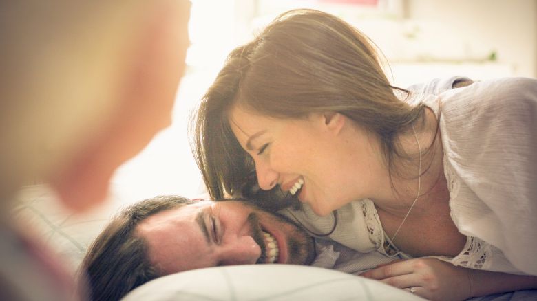 A woman laughing while lying on a man, both appearing happy and relaxed indoors with a third person partially visible in the foreground.