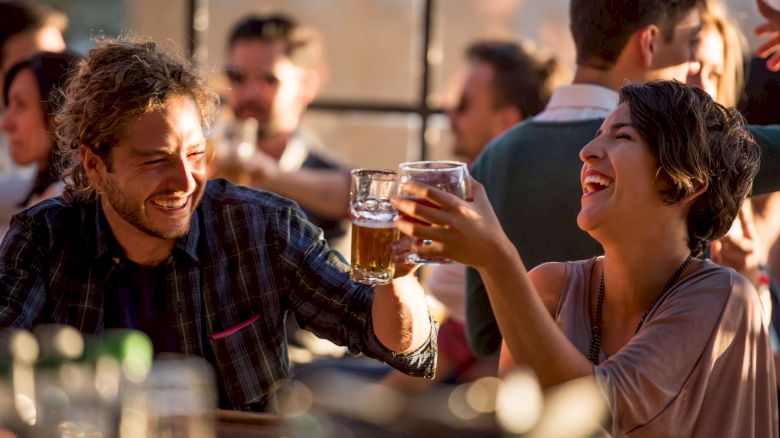 People are at a social gathering, smiling and clinking glasses, possibly enjoying drinks in a lively atmosphere.