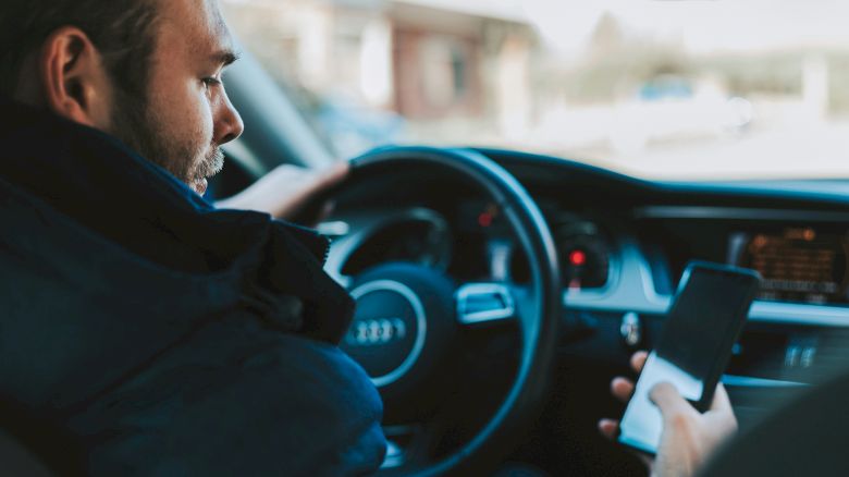 A person is sitting in a car, holding a smartphone while steering, with the car's dashboard and steering wheel visible.