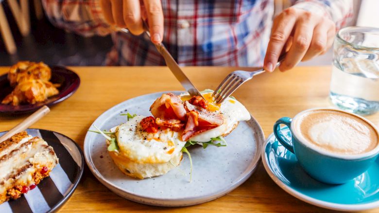 A person is cutting into a breakfast dish with eggs and bacon, accompanied by a cappuccino, a slice of cake, and a croissant.