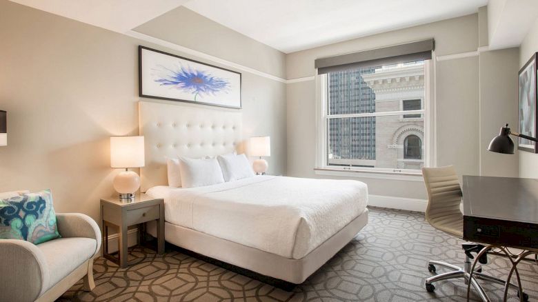 A modern hotel room with a bed, armchair, desk, and window view of a building, featuring neutral colors and abstract artwork above the bed.