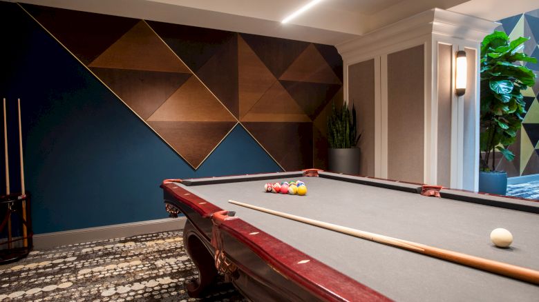 A modern room featuring a pool table, geometric wall art, and a potted plant, with cue sticks and pool balls set up for a game.