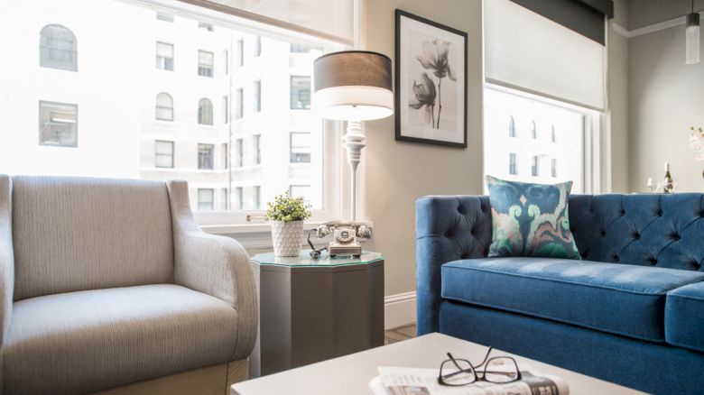 A cozy living room with a gray chair, blue sofa, lamp, framed art, and a table with glasses on a newspaper, near large windows.