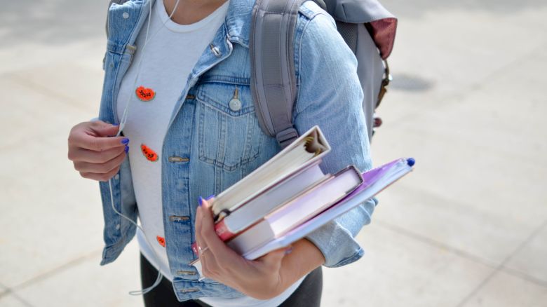 A person in a denim jacket carries books and a backpack, walking on a pavement.