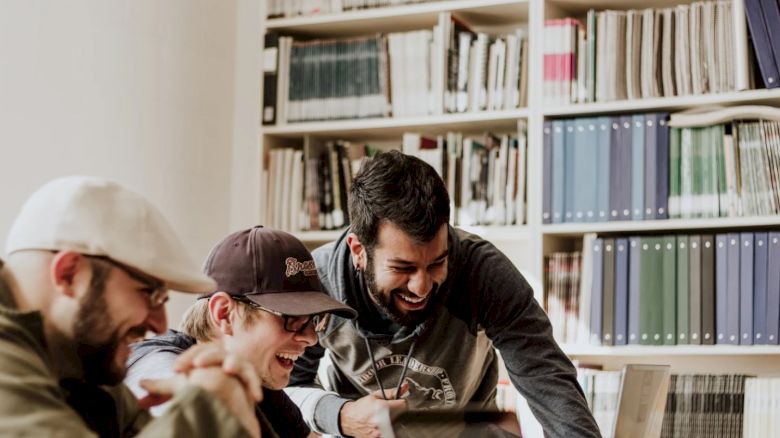 Three people are smiling and working at laptops in a library or office setting with bookshelves in the background.