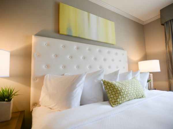 A neatly arranged bed with white pillows and a green decorative pillow, flanked by two lamps and a plant, with a yellow painting above.