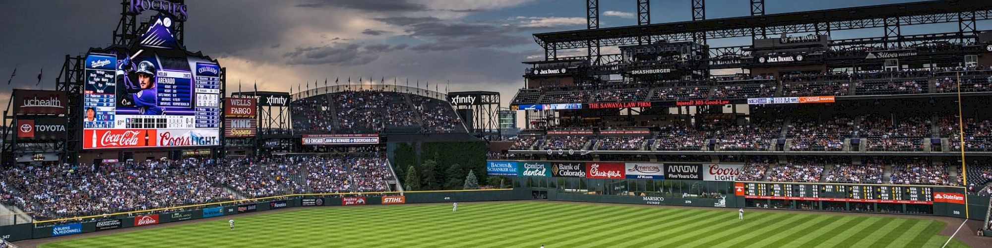 A baseball game is underway in a large stadium with spectators, featuring a big scoreboard and a cloudy sky overhead.