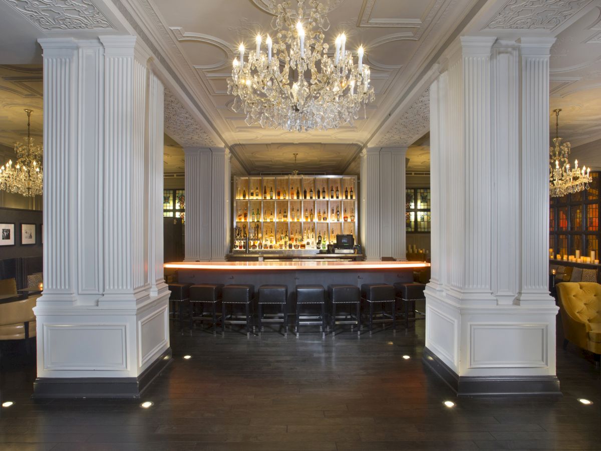 The image shows an elegant bar area with a chandelier, white columns, and a row of chairs in front of a well-stocked, illuminated bar.