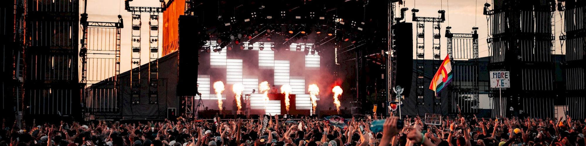 A large crowd is gathered in front of an illuminated outdoor concert stage during sunset, with some people raising their hands and a flag visible.