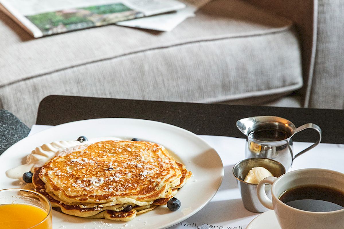 A cozy setting with pancakes, coffee, orange juice, and a newspaper on a table near a sofa.