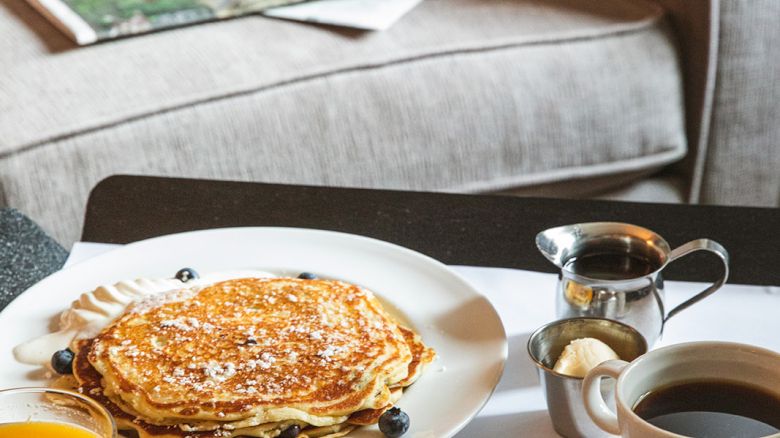 A cozy setting with pancakes, coffee, orange juice, and a newspaper on a table near a sofa.