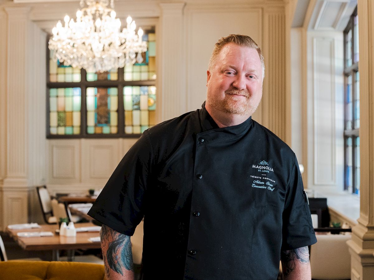 A chef in a black uniform stands in an elegant dining room with a stained glass window and chandelier, smiling at the camera.