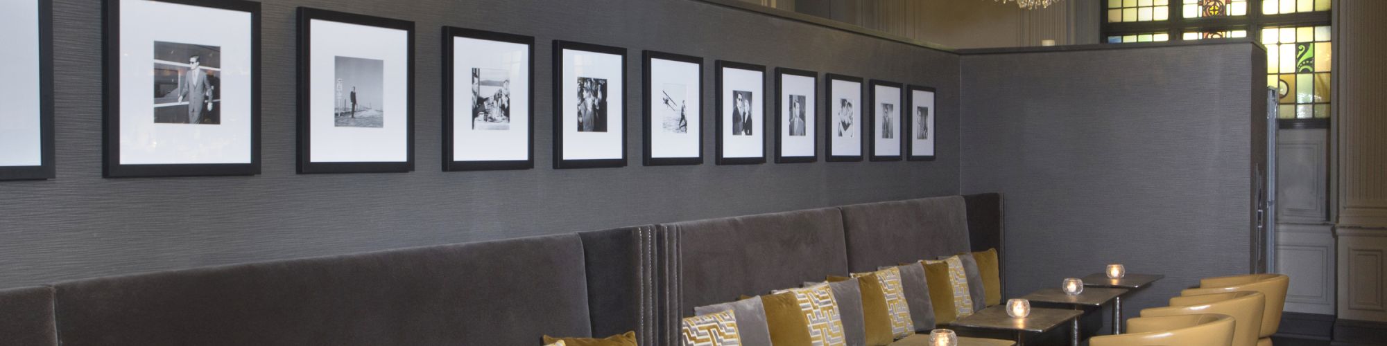 This image shows a stylish restaurant interior with chandeliers, framed photos, cushioned benches, and chairs arranged along dark tables.