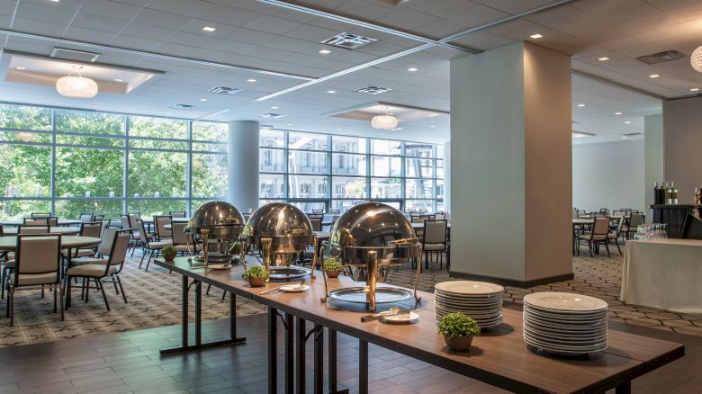 A modern buffet setup in a dining area with tables, chairs, and a chandelier. Large windows provide natural light, creating an inviting atmosphere.