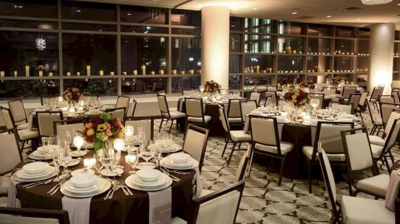 The image shows an elegant banquet hall with round tables set for a formal event. Candles, floral centerpieces, and place settings are on the tables.