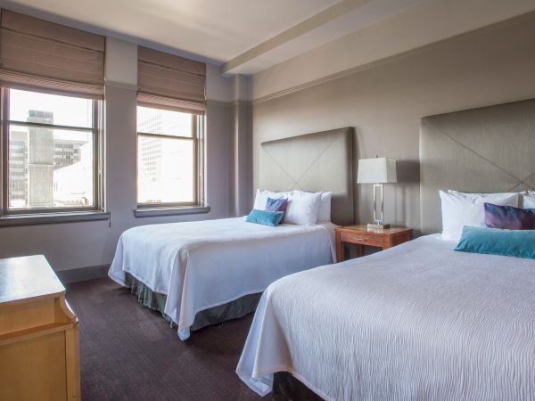 The image shows a hotel room with two double beds, a nightstand with a lamp, and a TV, featuring large windows and neutral decor.