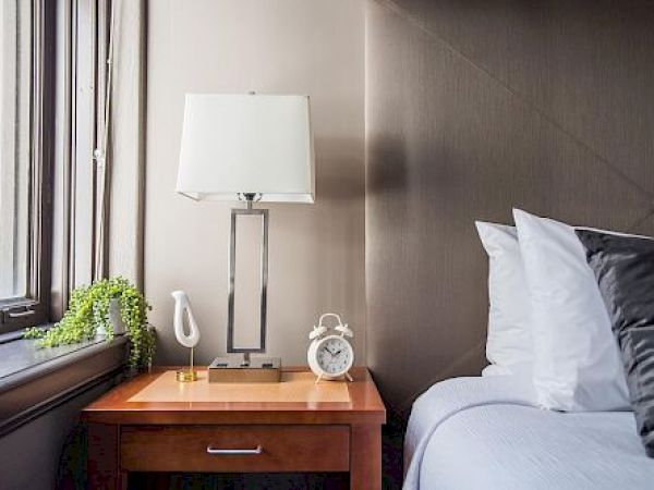 The image shows a bedroom with a bed, pillows, a side table, a lamp, a clock, and a small plant by the window.