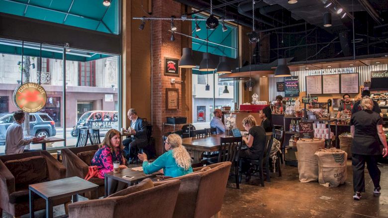 People are enjoying drinks and conversation in a cozy coffee shop with comfortable seating and large windows.