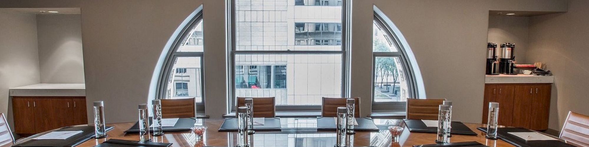 A modern conference room with a large oval table, chairs, notepads, and a chandelier. Two big windows show a city view outside.