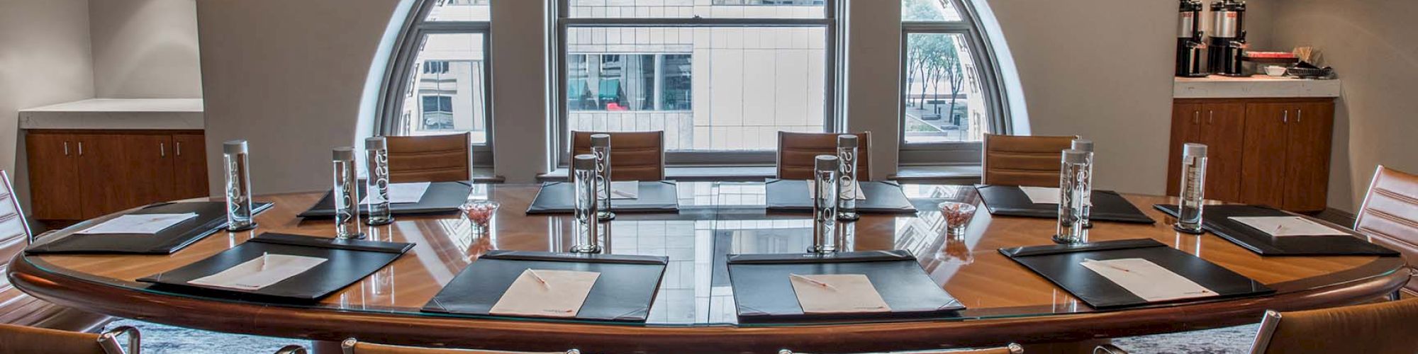 The image shows a modern conference room with a large oval table, beige chairs, a chandelier, and a city view through the windows.