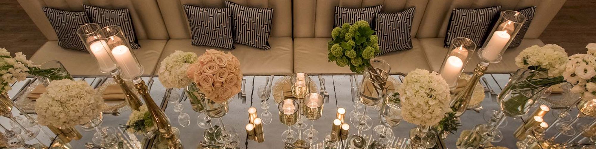 An elegantly set dining table with mirrored surfaces, floral centerpieces, lit candles, and cushioned seating with decorative pillows.