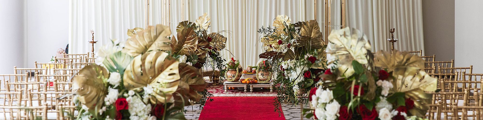 A beautifully decorated wedding venue with a gold and red theme, featuring floral arrangements, chairs, and a central aisle.