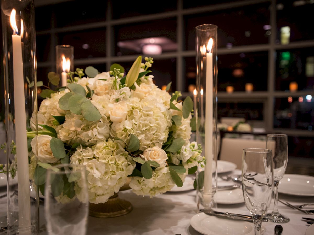 A table setting features a floral centerpiece with white roses and hydrangeas, surrounded by tall candles and elegant glassware.