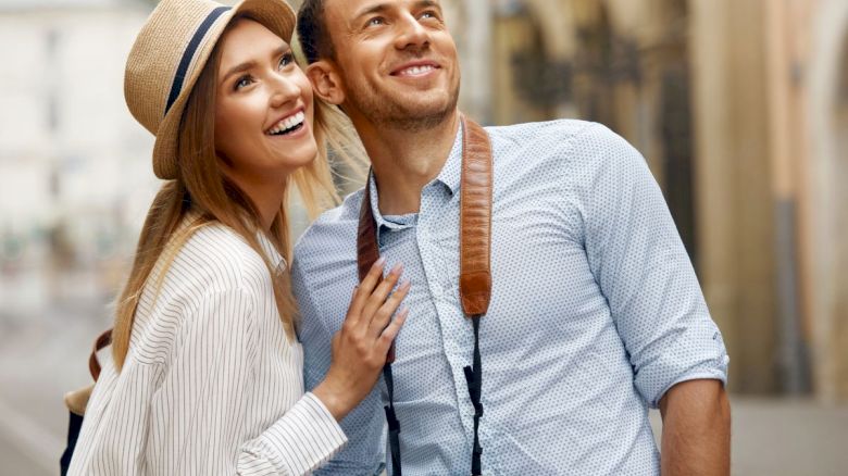 A couple smiling and looking upward, with the man holding a camera around his neck, strolling in a city setting.