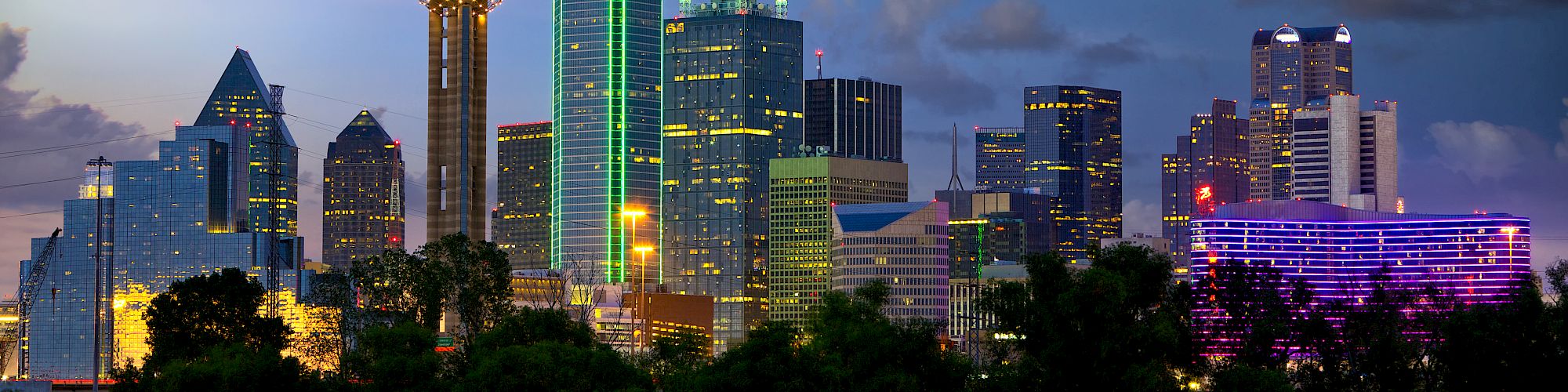 A vibrant city skyline at dusk with illuminated skyscrapers and iconic architecture under a partly cloudy sky.