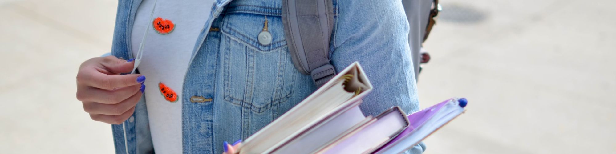 A person wearing a denim jacket and carrying a backpack is holding books while walking outside.