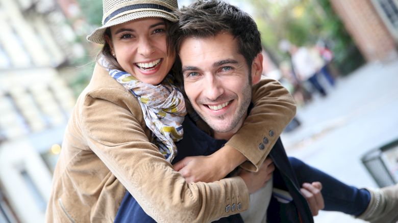 A smiling couple outside; the woman piggybacks on the man, both looking happy and casual in a relaxed, urban setting.