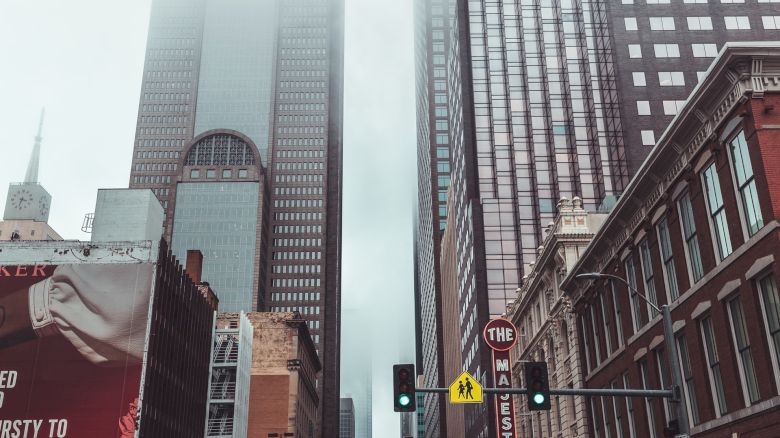 A city street with skyscrapers obscured by fog, wet pavement, traffic lights, and billboards. Vehicles and pedestrians are visible.