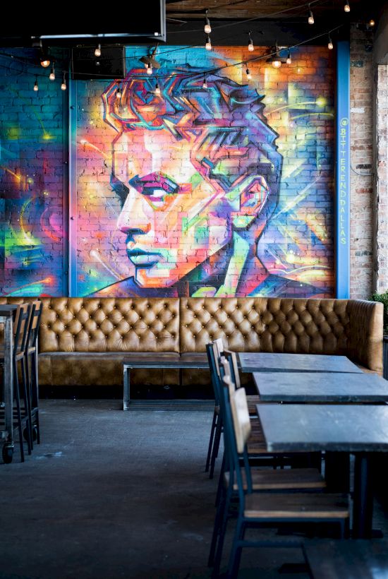 A colorful mural of a stylized face on a brick wall above a tan leather sofa, with tables and chairs in the foreground, in a café setting.