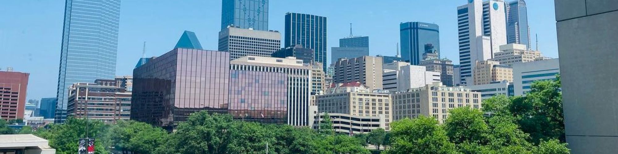 A city skyline with various skyscrapers, a clear blue sky, and a few trees along the street in the foreground.