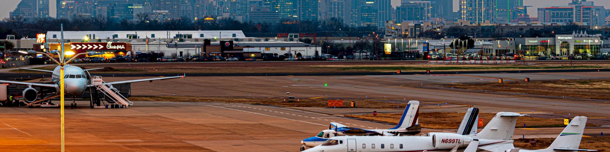 An airport with several private jets and a commercial plane on the tarmac, with a city skyline in the background at dusk.