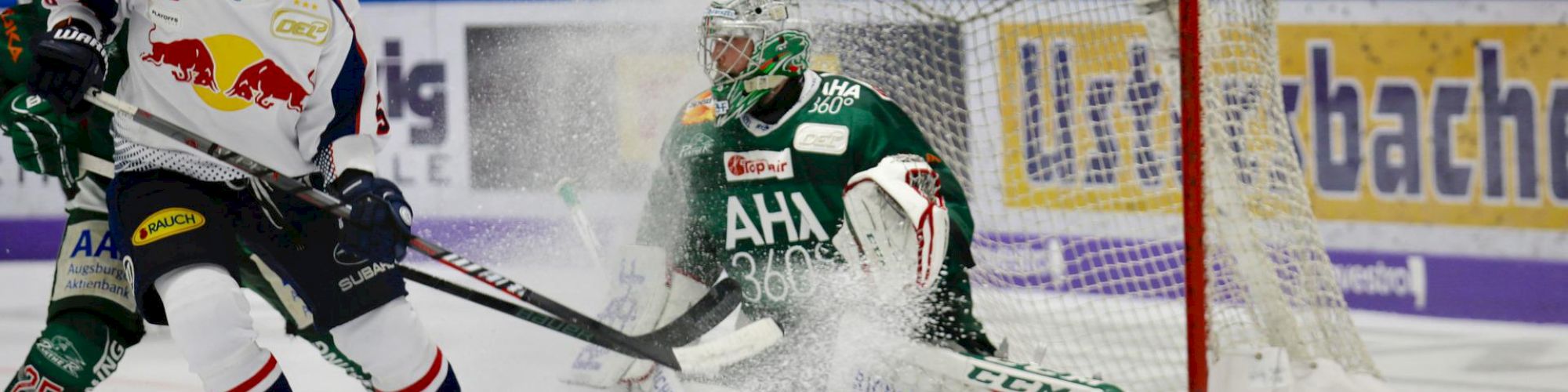 Hockey players in action; one shoots the puck as the goalie defends the net, with ice spray from the intense play.