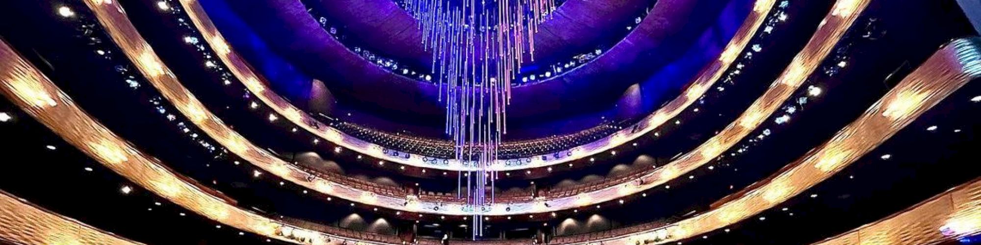 It's an empty modern theater with tiered seating and a striking blue-lit ceiling chandelier, viewed from the stage.