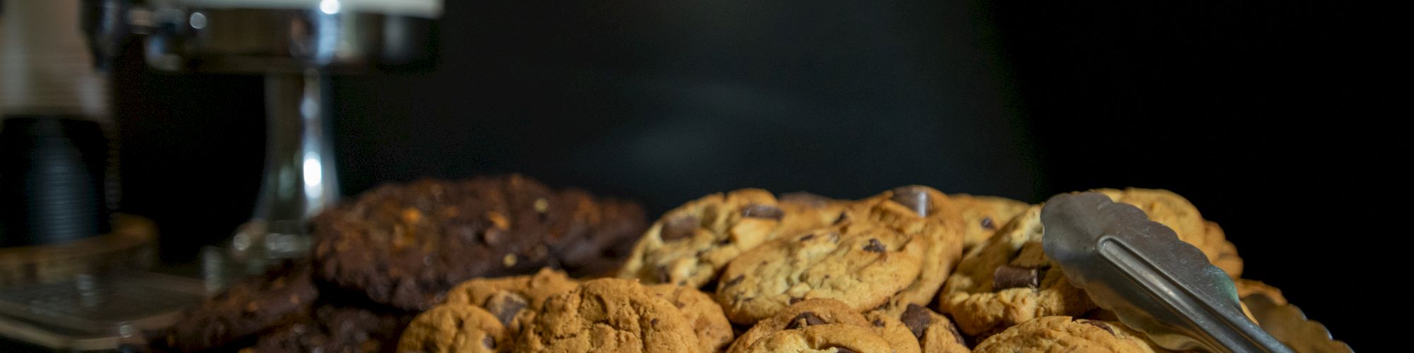 A wooden tray with chocolate chip cookies, tongs, and a glass container of milk in the background ends the setup.