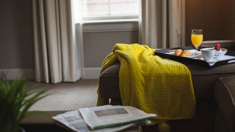 A cozy room with a yellow blanket on a chair, breakfast tray with juice and pastries, and a newspaper on the armrest near a window.