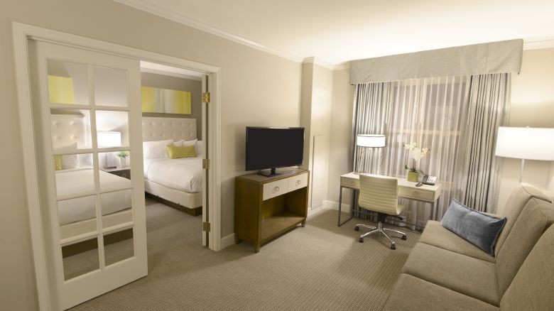 The image shows a hotel suite with a bed, glass doors, a TV, a desk with a chair, and a couch with a lamp in a modern, neutral design.