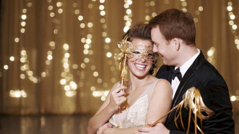 A couple in formal wear enjoys a masquerade, holding masks in front of twinkling lights. They share a joyful, intimate moment together.