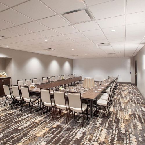 A modern conference room with a long table, surrounded by chairs, neutral decor, and patterned carpet, designed for meetings or events.