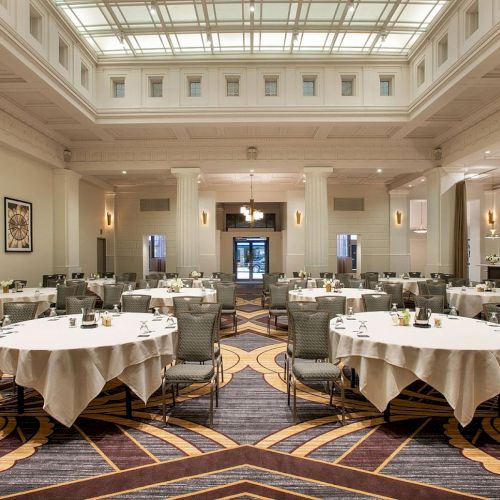 The image shows an elegant banquet hall with round tables set for dining under a high ceiling and large windows.