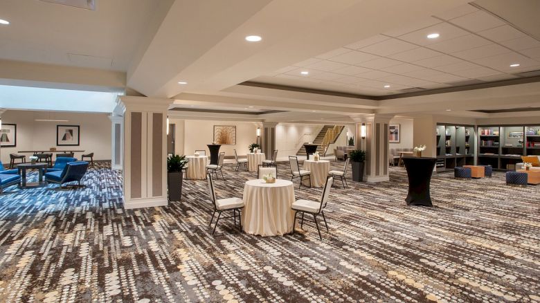 The image shows a spacious room with round tables and chairs, elegant carpeting, and modern decor, likely set up for an event.