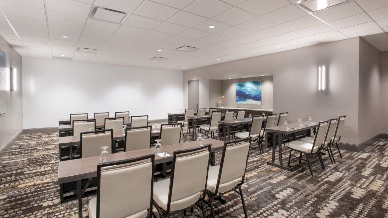 The image shows a modern conference room with rows of chairs and tables, a patterned carpet, and a painting on the wall.