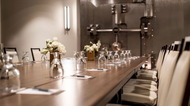 The image shows a long conference table set with upside-down glasses, notepads, and flowers, in a room with industrial decor elements.