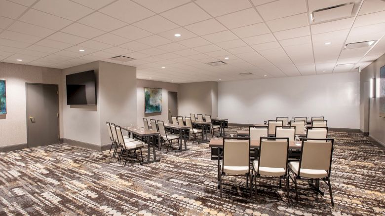 The image shows a conference room with rows of chairs and tables on a patterned carpet. There's a TV on the wall and framed artwork.